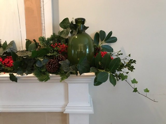 White mantel with holly, ivy, pinecones, and a green glass jug — holiday decorating by Christopher Patrick Interiors